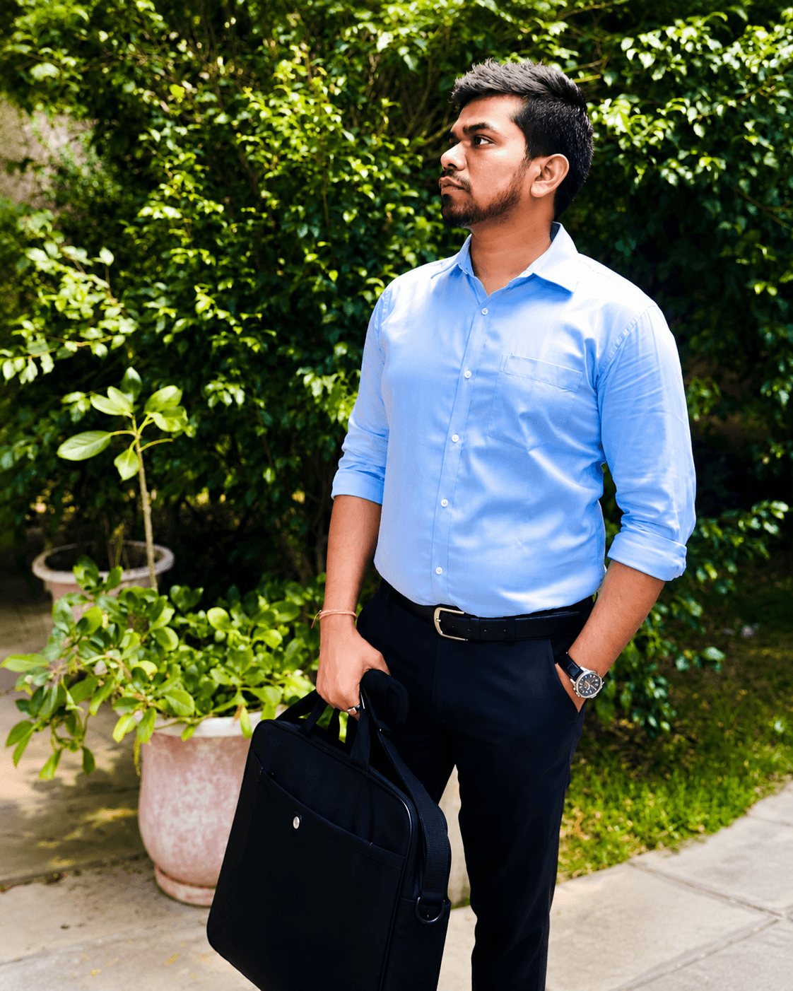 Manas Majhi walking through a garden path holding a laptop bag, looking off to the side
