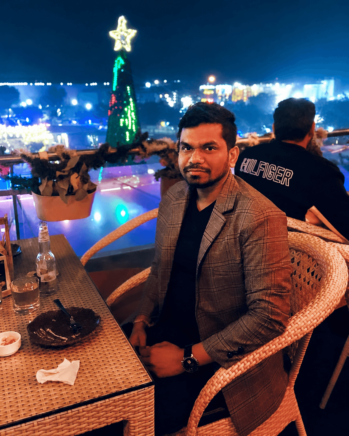 Manas Majhi on a rooftop terrace at night, city skyline and lit Christmas tree in the background